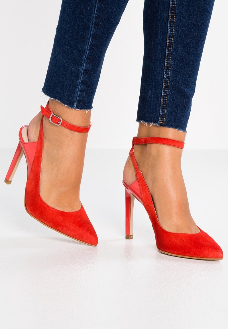 Pieds portant des escarpins en daim rouge vif avec des brides à la cheville, assortis à un jean bleu foncé raccourci sur une surface blanche.