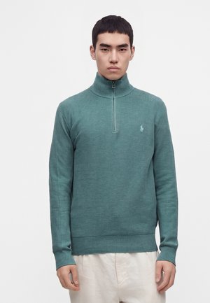 Mand iført en teal halv-lynlås trøje med et lille logo på brystet og lyse bukser, der står mod en ensfarvet baggrund.