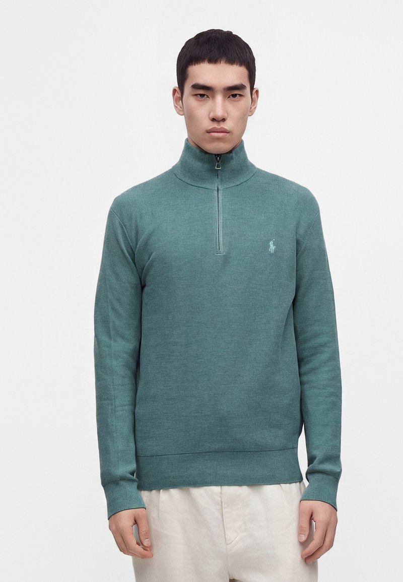 Homme portant un pull demi-fermeture éclair couleur sarcelle avec un petit logo sur la poitrine et un pantalon clair, debout devant un fond uni.