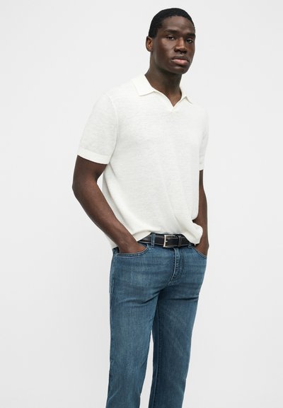 Άνδρας που φοράει λευκό πόλο μπλουζάκι με κοντά μανίκια, μπαίνει μέσα σε μπλε τζιν με μαύρη ζώνη, στέκεται με τα χέρια στις τσέπες.