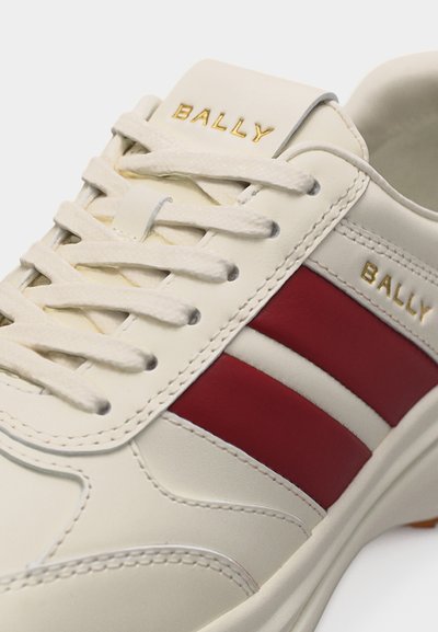 Baskets en cuir blanc avec des rayures rouges, des lacets blancs, et "BALLY" embossé en or sur la languette et le panneau latéral.