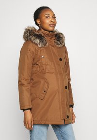 Parka marrón con capucha de piel sintética, botones frontales, bolsillos laterales y cintura elástica. Usada sobre jeans azules. Textura suave.