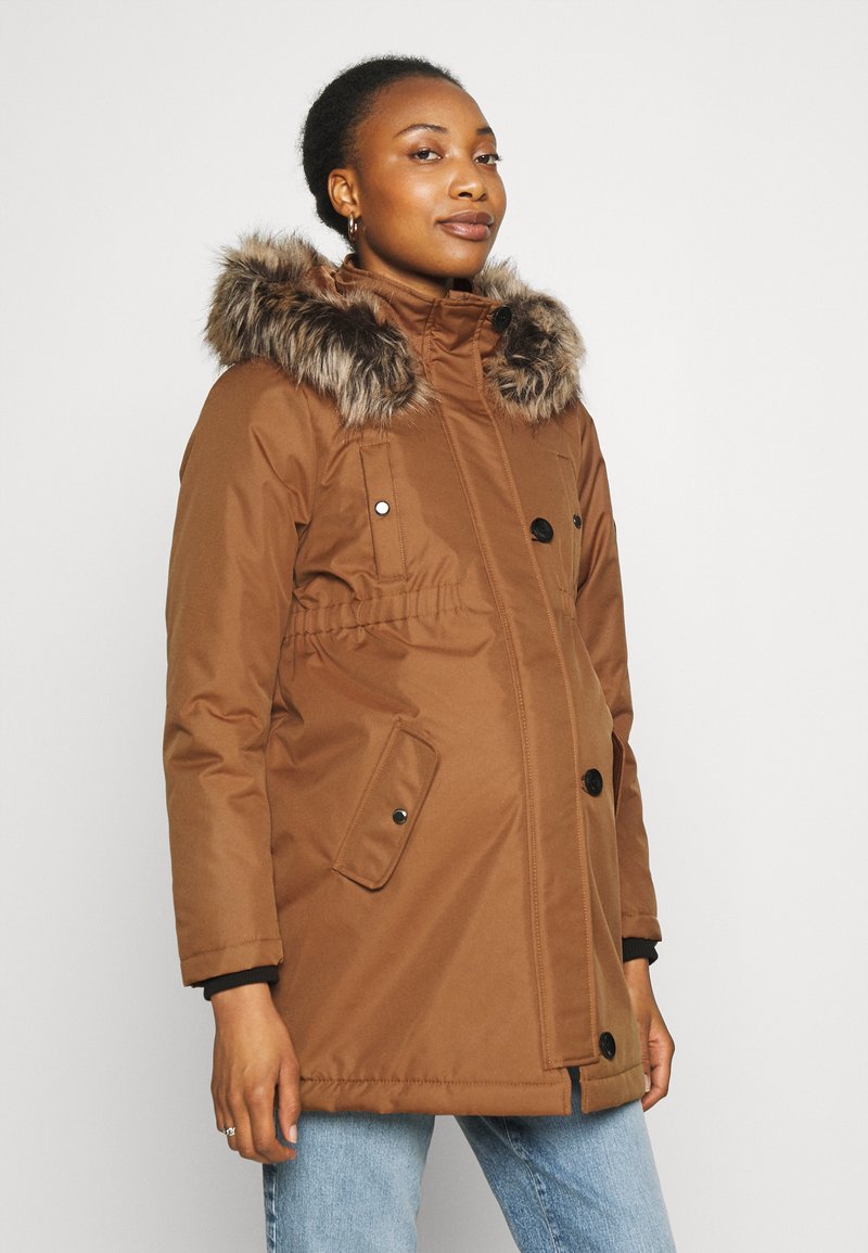 Parka marrón con capucha de piel sintética, botones frontales, bolsillos laterales y cintura elástica. Usada sobre jeans azules. Textura suave.