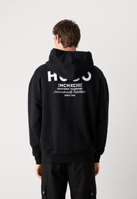 Svart hettegenser med et stort hvitt logo på ryggen, ribbede mansjetter og kant. Myk tekstur, med snorhette.
