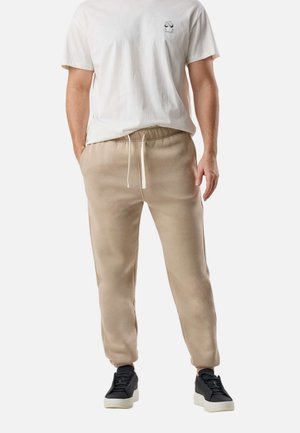 Hvid T-shirt med korte ærmer og en lille grafik på brystet, beige sweatpants med snøre i taljen og sorte sneakers med hvide såler.