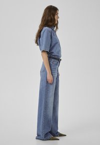 Ženska z dolgimi rjavimi lasmi stoji v profilu, oblečena v ohlapno denim srajco, kavbojke z visokim pasom in širokimi hlačnicami, črn pas in čevlje z ostrim prstom.