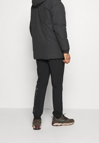 Uomo con giacca nera, pantaloni neri con il logo "Columbia" e scarpe da trekking marroni, in piedi contro una parete chiara e semplice.