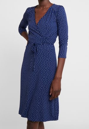 Kobieta w granatowej sukience do kolan w białe kropki, z rękawami do połowy przedramienia, dekoltem w serek i paskiem wiązanym w talii.