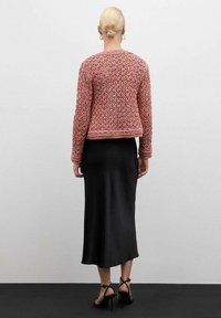 Růžová pletená bunda s otevřen�ým vzorem, doplněná o přiléhavou černou saténovou sukni a černé sandály na podpatku. Pohled zpoza.