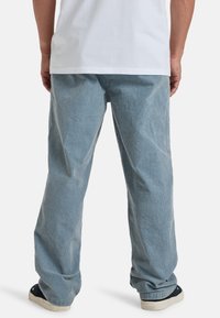 Pantalons en velours côtelé bleu clair avec une coupe décontractée, présentant un design à jambe droite et une surface texturée, associés à des chaussures sombres.