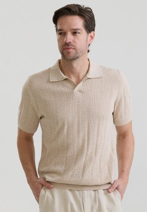 Mand med brunt hår iført en beige kortærmet strikket poloskjorte og lysebeige bukser, stående med hænderne i lommerne.