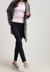 Cappotto di lana grigio, maglione rosa chiaro, pantaloni slim-fit blu scuro e scarpe slip-on grigie con suole bianche, che mostrano un design minimalista.