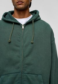 Donkergroen hoodie met rits en trekkoord, grote voorzakken en een gestructureerde afwerking. Voorzien van een metalen rits en een gelaagd wit T-shirt eronder.