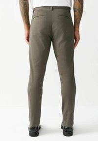 Slim-fit olive green trousers with a smooth texture, two back pockets, and a flat waistband. Paired with black shoes and a white shirt.
