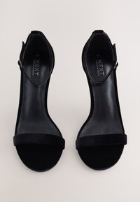 Crne sandale s visokim petama od baršunaste tkanine, s remenčićem oko gležnja i glatkom, sjajnom unutarnjom podstavom. Ukrasni logo unutra.