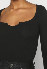 Czarny, prążkowany top z długim rękawem, z dekoltem w serek i delikatnym wycięciem w kształcie litery V. Połączony z czarnymi, skórzanymi spodniami z wysokim stanem.