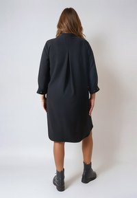 Zwarte jurk met een kraag, lange mouwen en een rugplooi. Gestyled met zwarte enkellaarsjes met een gladde textuur.