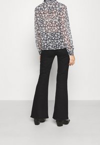 Personne portant un pantalon noir évasé, des chaussures à talons noires et un chemisier transparent à manches longues à motif floral, se tenant devant un fond blanc uni.