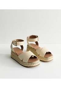 Beige kilklackssandaler med korsremmar, tillverkade av slät syntetisk material, med en texturerad juteplattform som accentueras av gyllene detaljer.