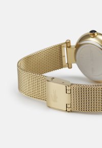 Bracelet de montre en maille dorée avec une boucle métallique portant un logo. Le boîtier de la montre est rond avec une petite pierre bleue au niveau de la couronne.
