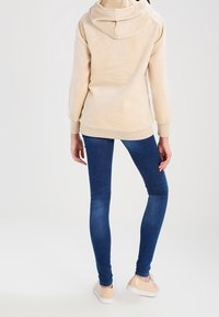 Sweat à capuche beige en polaire avec capuche, manches longues et poignets côtelés, associé à un jean skinny bleu foncé et des baskets de couleur claire.