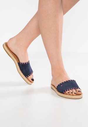 Feet wearing navy blue slide sandals with woven rope sole and red, white, and blue trim on a white background.
