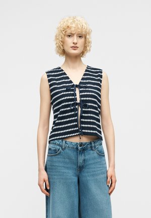 Göndör szőke hajú nő ujjatlan, sötétkék-fehér csíkos, elöl megkötött felsőben és magasderekú, bőszárú kék farmert visel, egyszerű háttér előtt.