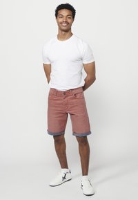 Shorts en denim rouge avec des ourlets roulés, associés à un t-shirt blanc uni et des baskets blanches avec des accents noirs. Fond neutre.