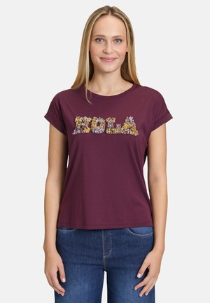 T-shirt bordeaux avec des manches courtes roulées et un texte floral "Hola" en jaune et violet. Matière en coton douce et coupe décontractée.