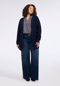 Cardigan blu navy con dettagli scintillanti, abbinato a una camicetta con motivo geometrico e jeans blu a gamba larga. Scarpe nere completano il look.