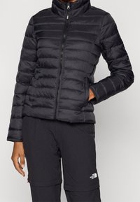 Sort pufferjakke med høj krave, lynlås fortil og quiltede paneler. Har et slankt materiale og sidelommer. Bæres med sorte bukser.