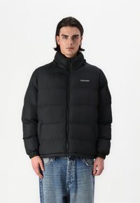 Μαύρο τζάκετ πούφ φτιαγμένο με ψηλό γιακά, κλείσιμο με φερμουάρ και ελαστικούς καρπούς. Διαθέτει λογότυπο στην αριστερή πλευρά του στήθους και απαλό υλικό.