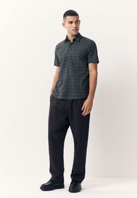 Crna košulja kratkih rukava s teksturiranim uzorkom, uparena s širokim tamnim hlačama i crnim cipelama, tvoreći ležernu odjeću.