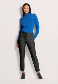 Vrouw die tegen een muur staat, gekleed in een blauwe turtlenecktrui, zwarte taps toelopende broek met trekkoord en zwarte sneakers.