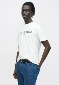 Noor mees punutud juustega, kandes valget Calvin Kleini t-särki, siniseid teksaseid ja musta vööd, seisab ühe käe taskus.