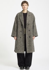 Dobbeltradet frakke i struktureret sort-hvid hørslagsmønster, med store revers og fire knapper. Båret over en mørk strikket sweater.