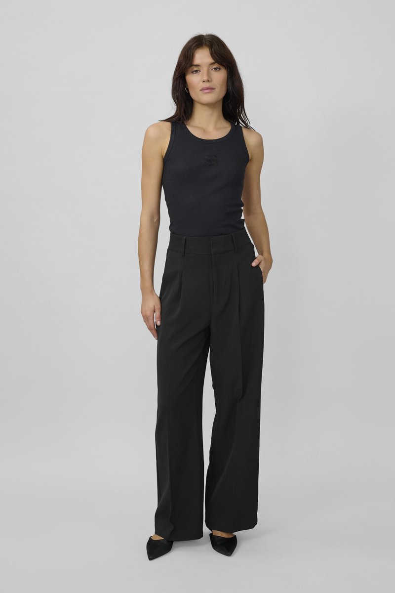 Black ribbed tank top with a subtle logo, paired with high-waisted black pleated trousers. Model stands with one hand in pocket.