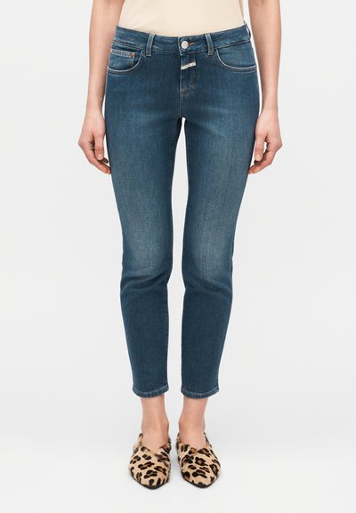 Jean slim bleu foncé avec légère décoloration, cinq poches et fermeture à bouton sur le devant, associé à des chaussures slip-on imprimé léopard.