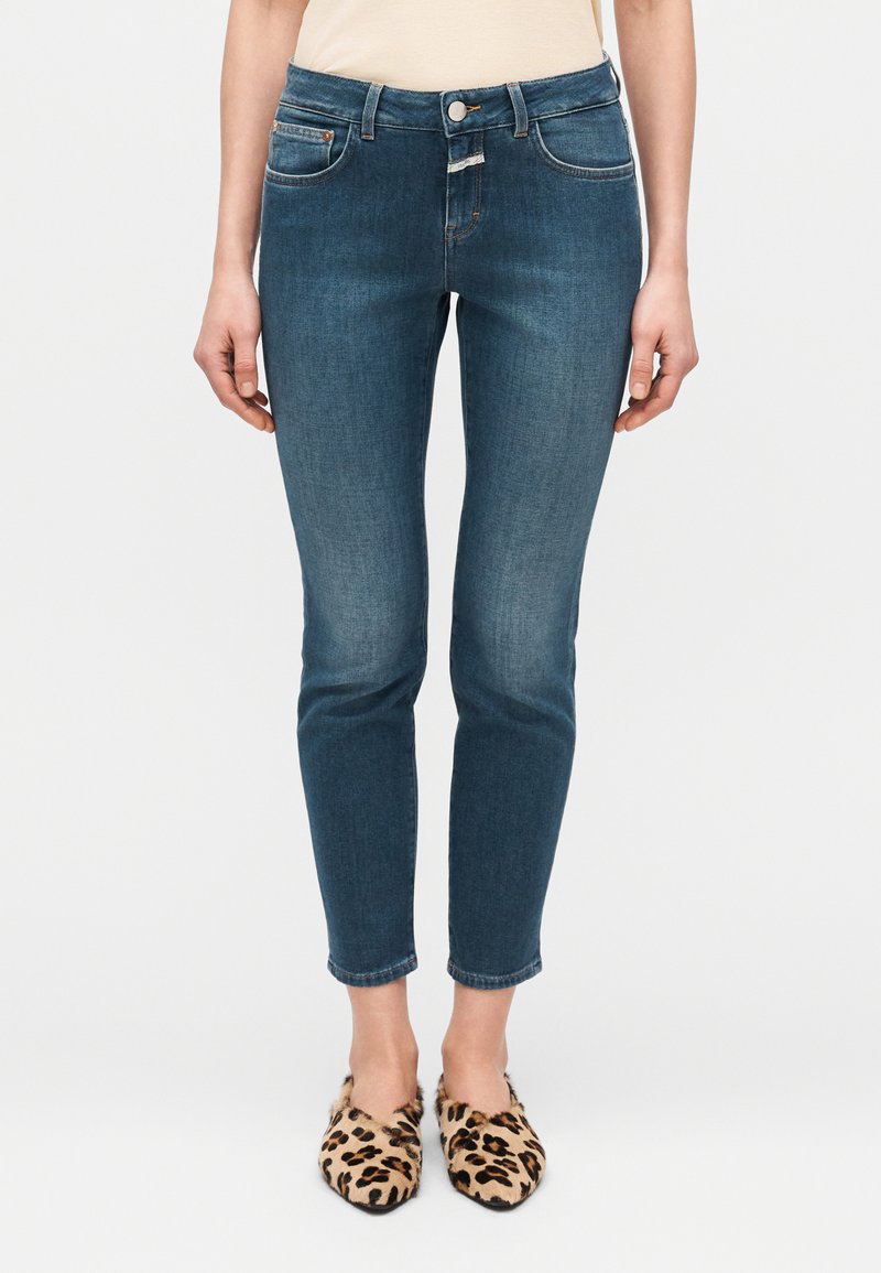 Jeans blu scuro a taglio slim con delicato effetto sbiadito, cinque tasche e chiusura frontale con bottoni, abbinati a scarpe slip-on con stampa leopardata.