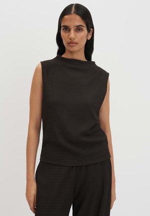 Femme aux longs cheveux foncés portant un haut noir texturé sans manches et un pantalon noir assorti, debout devant un fond clair uni.