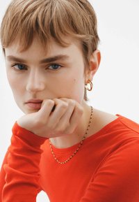 Pendiente de aro dorado, collar delicado con pequeñas cuentas, blusa roja de manga larga. Textura suave, fondo neutro, expresión facial concentrada.