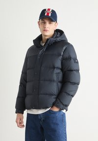 Giubbotto imbottito blu navy con cappuccio, caratterizzato da grandi sezioni trapuntate e chiusure a scatto. Accompagnato da un berretto blu navy con un logo rosso e bianco.