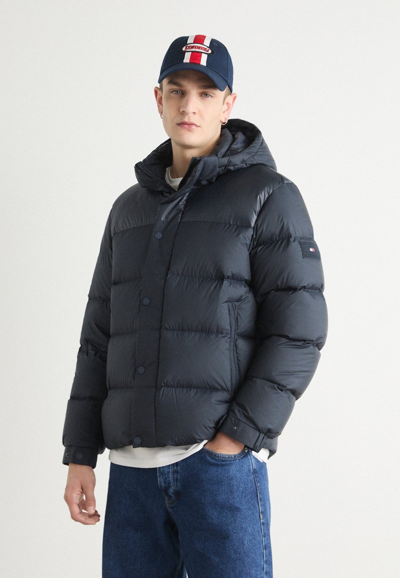 Giubbotto imbottito blu navy con cappuccio, caratterizzato da grandi sezioni trapuntate e chiusure a scatto. Accompagnato da un berretto blu navy con un logo rosso e bianco.