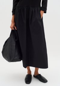 Schwarzes Kleid mit gerafftem Bund, aus einem glatten Stoff gefertigt. Kombiniert mit schwarzen Leder-Mules und einer strukturierten schwarzen Tote-Tasche.