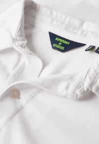 Weißes Baumwoll-Poloshirt mit einem Knopfkragen, versehen mit einem blau-grünen Markenetikett "SUPERDRY @ STUDIOS", das gut sichtbar im Inneren angebracht ist.
