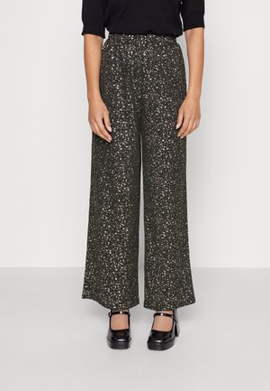 Femme portant un pantalon noir taille haute, à jambes larges, avec un motif tacheté blanc et des talons Mary Jane noirs, debout devant un fond clair uni.