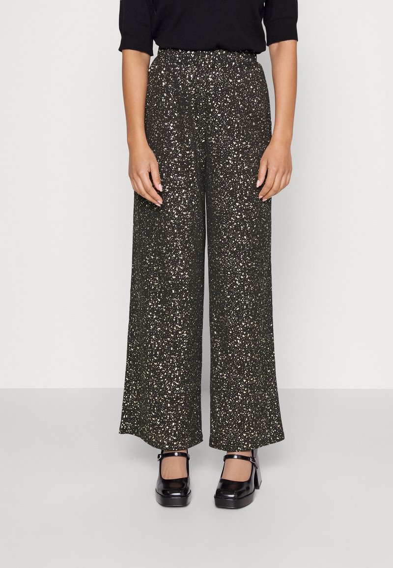 Femme portant un pantalon noir taille haute, à jambes larges, avec un motif tacheté blanc et des talons Mary Jane noirs, debout devant un fond clair uni.