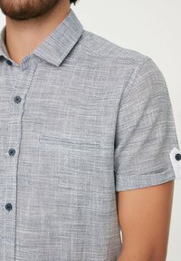 Camisa de botones de manga corta en una tela texturizada azul y blanca, con un bolsillo en el pecho y botones oscuros.