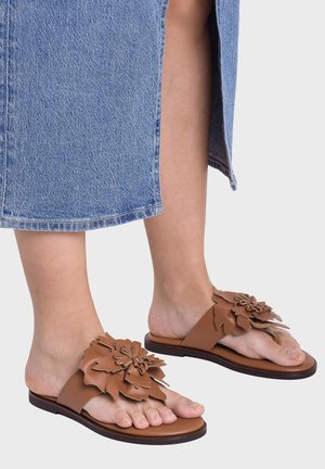 Stopy w brązowych skórzanych sandałach wsuwanych z wielowarstwowymi ozdobami kwiatowymi, zestawione z niebieską jeansową spódnicą.