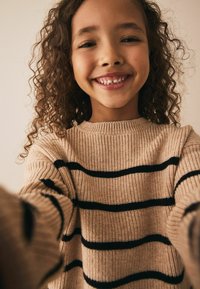 Strikket sweater i beige med sorte vandrette striber, der har en ribbede halsudskæring og struktureret stof. Nærbillede af et smilende barn iført den.
